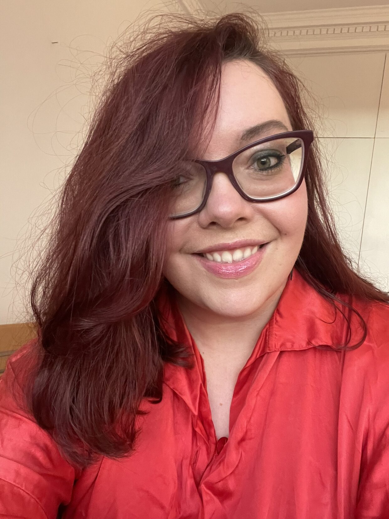 Selfie of Jess wearing a red blouse. Jess has long brown hair, thick-framed glasses and is smiling at the camera