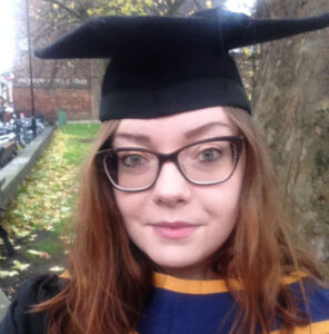 Selfie of Jess wearing graduation cap and gown. Jess has long brown hair and is wearing thick-framed glasses