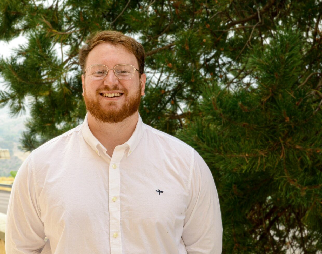 Daniel Fleming has short red hair, glasses and a beard. Daniel is wearing a white button down shirt and is standing next to a tree in Utah