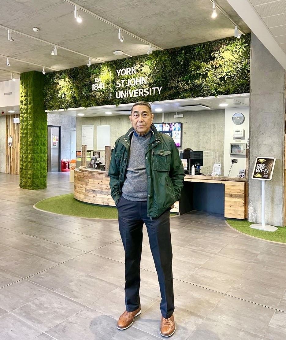 Noke wears dark trousers, a grey top and a dark waterproof jacket. Noke has short hair. Noke is standing in front of York St John University's reception area.