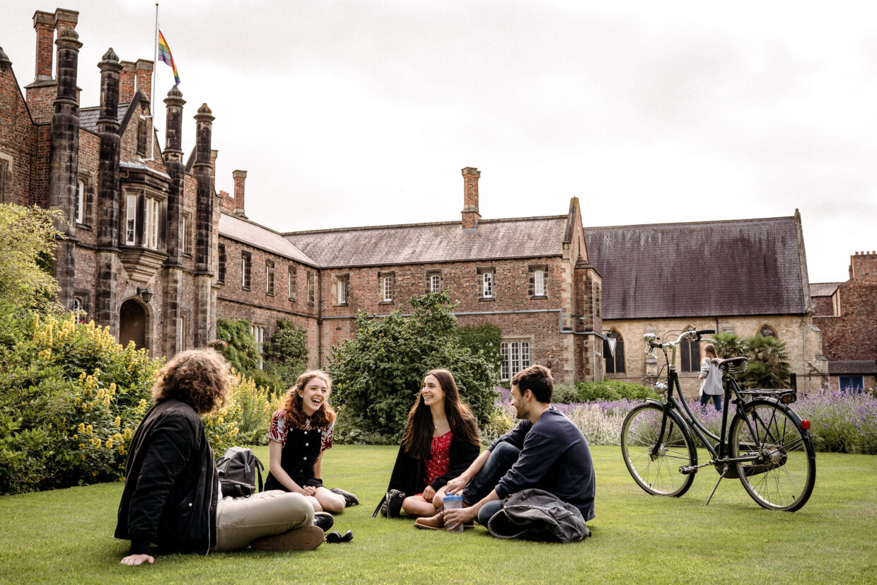 A side view of the main Lord Mayor's Walk campus with the pride flag flying and four students sitting on the grass smiling. There is a bike propped up on its stand next to the students.
