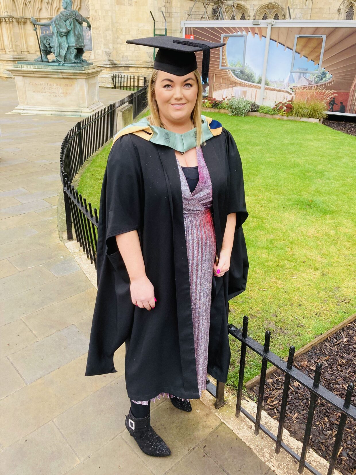 Chloe wears a graduation cap and gown and is stood outside of York Minster. Chloe has shoulder length blonde hair and is wearing a pink dress.