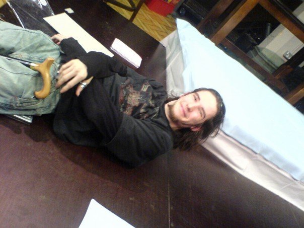 Picture of Dom laying on the floor next to a mattress as a student. Dom has long brown hair and it wearing a long sleeved black top and dark tshirt