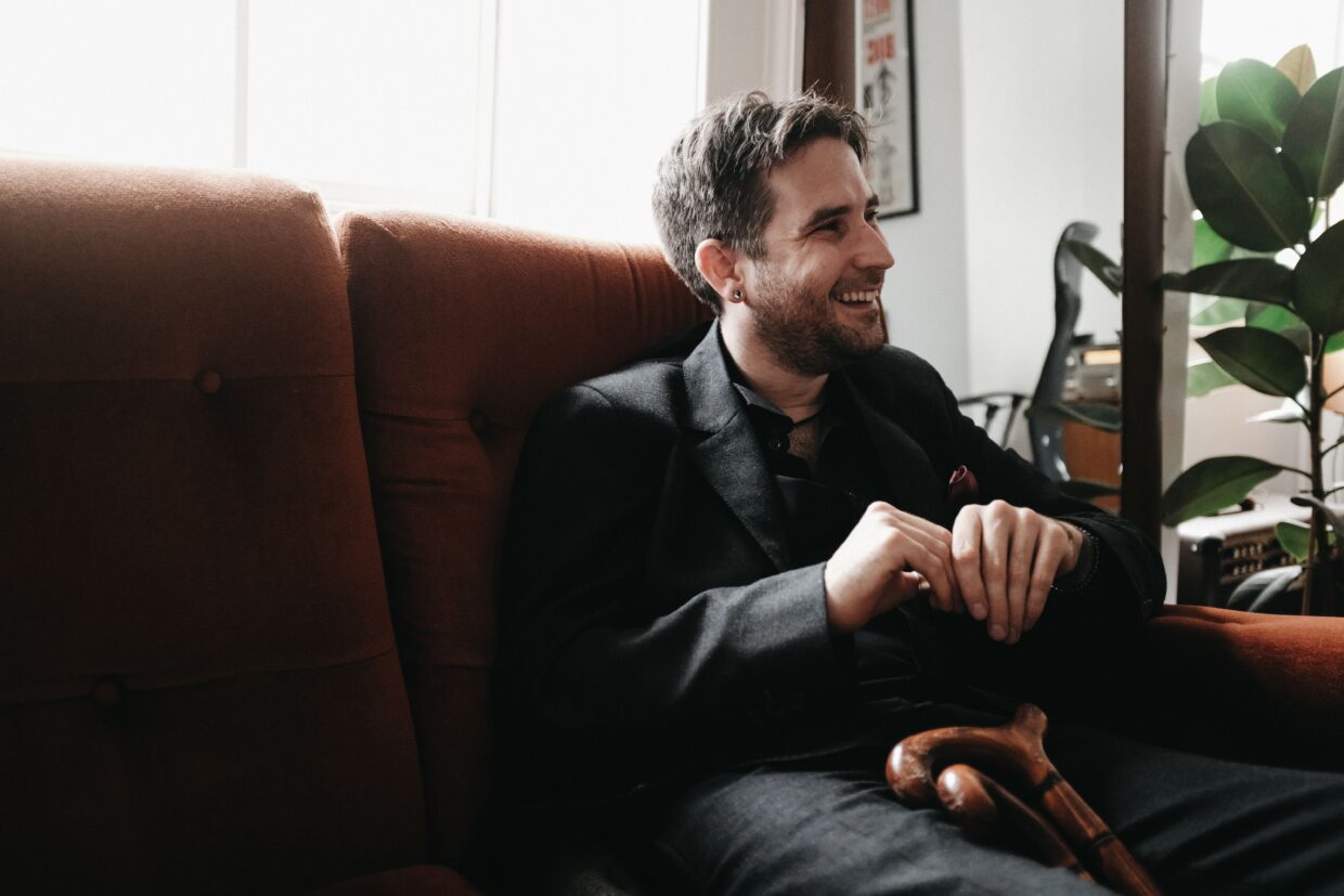 Dom Smith has short brown hair and is wearing a blazer and dark shirt. He is sitting on a sofa looking away from the camera