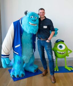 A picture of Nathan in San Francisco standing with Mike and Sully from Monsters Inc at Pixar Studios. Nathan has short hair and is wearing a blue shirt, blue jeans and brown shoes.