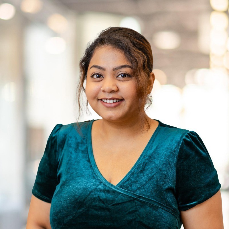 Farah has brown hair tied in a bun. Farah is wearing a teal velvet top and is smiling at the camera.