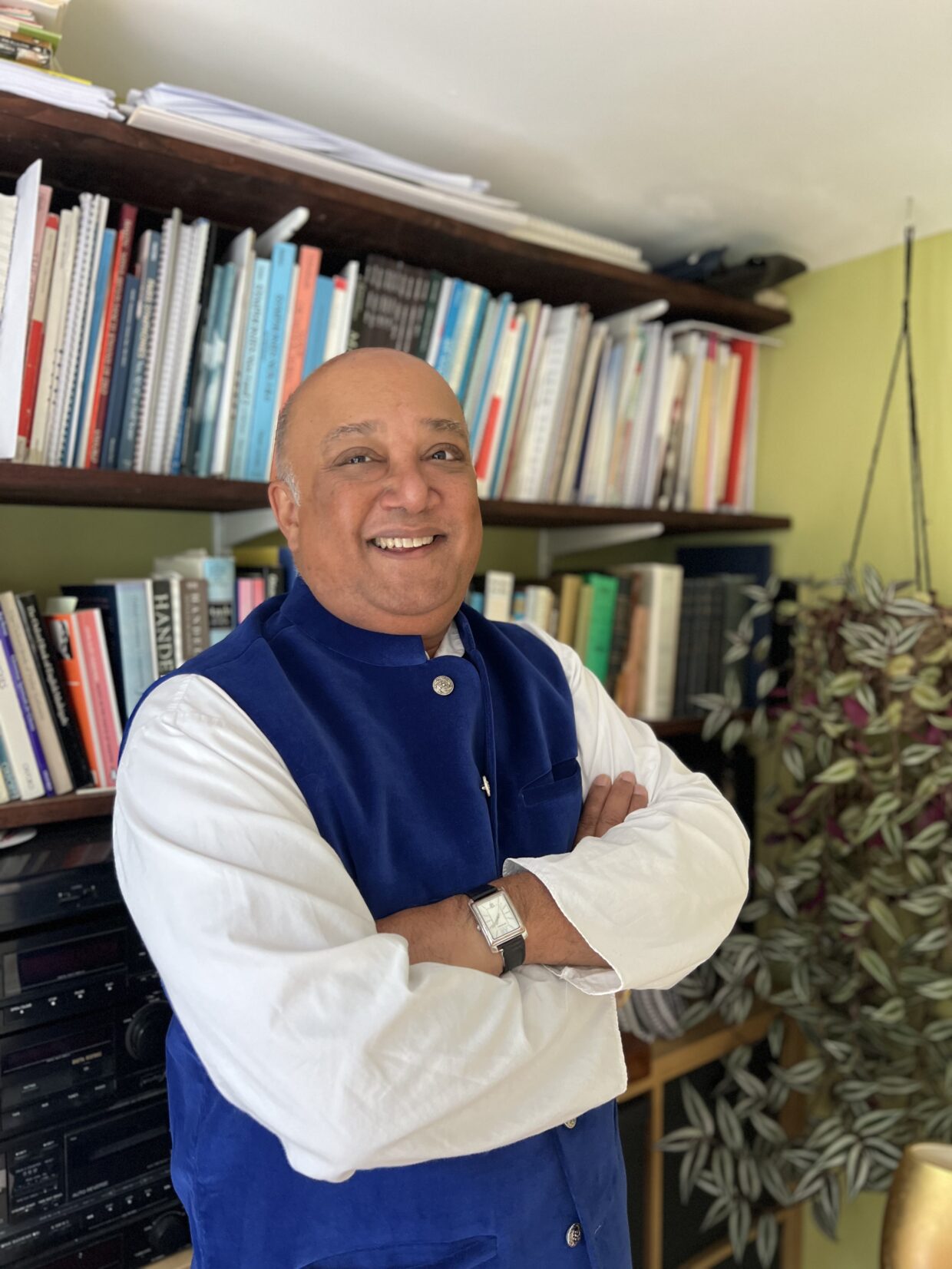 Manvinder has very short grey hair and it smiling at the camera. Manvinder is stood in front of a bookshelf, with his arms folded, and wearing a navy blue and white top.