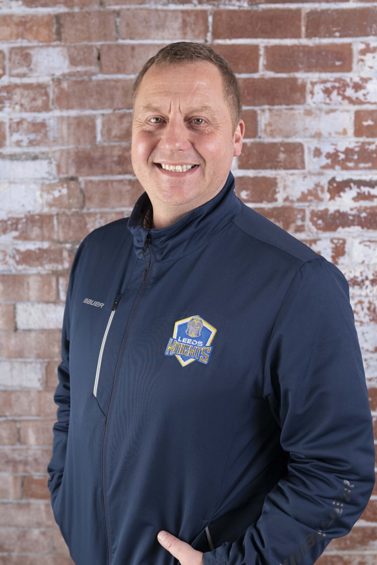 Warwick has short brown hair and is smiling at the camera. Warwick is wearing a navy sports fleece with a Leeds Knights logo