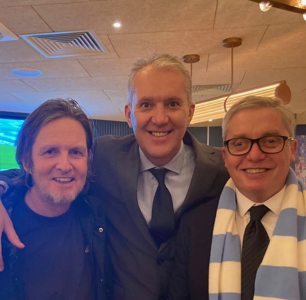 3 men stand posed for a photo. David is in the middle and has short grey hair and is wearing a suit. David's arms are around his friends Chris and Simon who he met on his first day of York St John