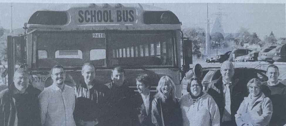 Black and white image of people stood in front of a school bus. John is on the far left hand side.