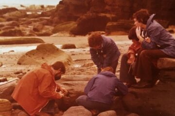 A slightly yellow tinged image, it shows a group of students on the beach looking in rock pools and at rocks. They are all wearing waterproofs.