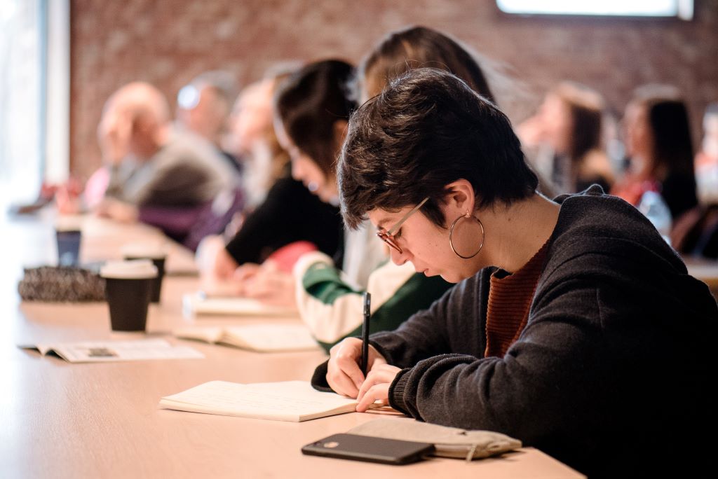 A woman taking notes at a long desk during a conference.