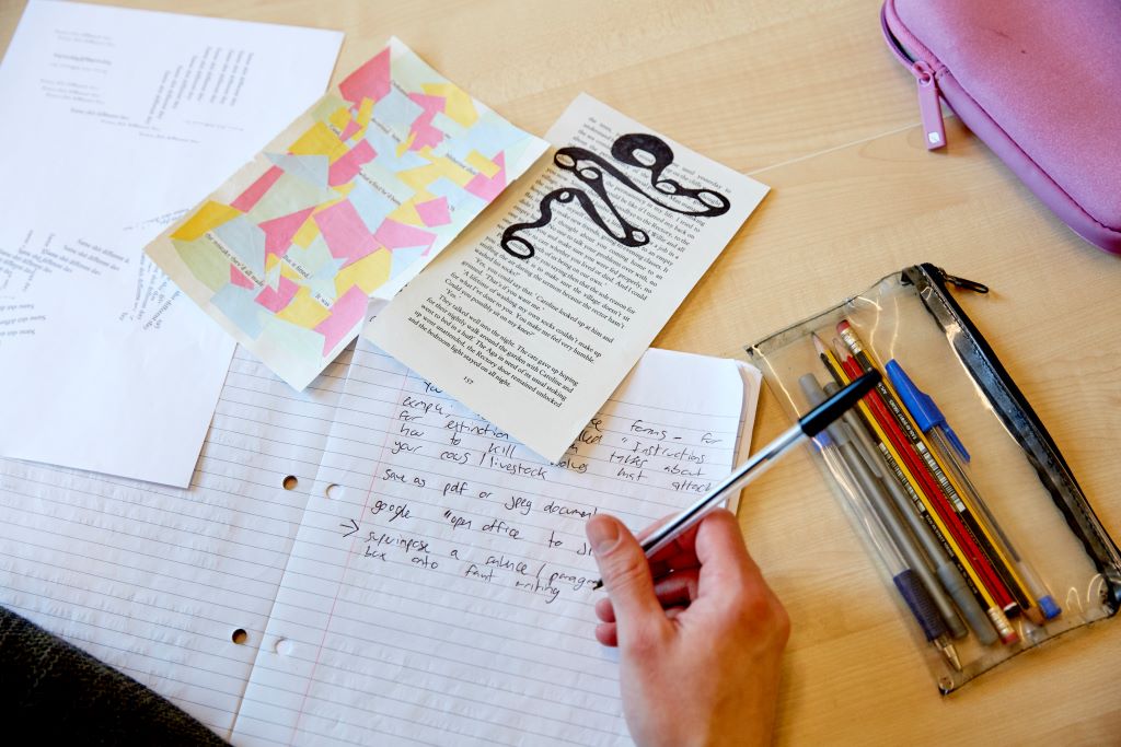 A table with a pencil case, scrap paper and a notepad in use.