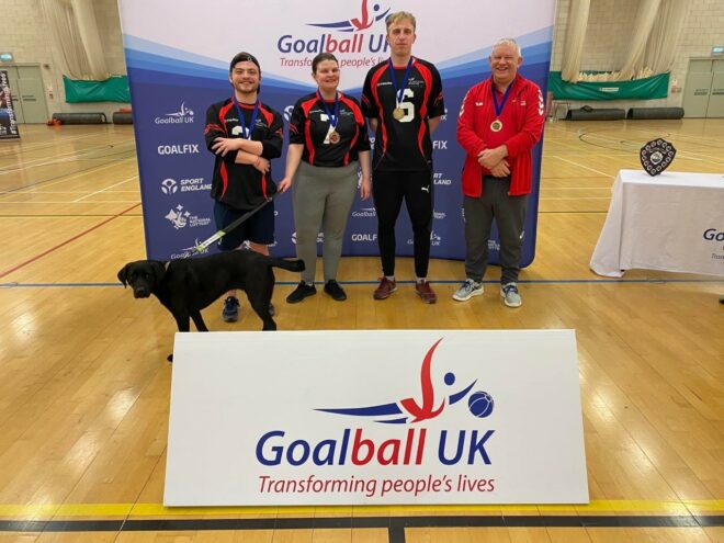 The image contains four people, and one guide dog. The people are dressed in sports gear, behind a sign that reads 'Goalball UK'