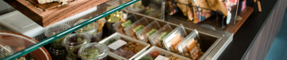 A photo of a cool box inset into the counter of a café, containing sandwiches, salad and pasta pots. there is a wooden shelf above containing brownie slices with different chocolate chips